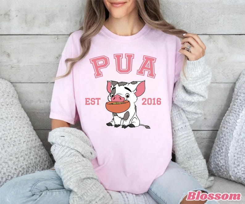 May include: Light pink t-shirt featuring the word "PUA" in pink block letters above a cartoon pig holding a bowl of food. The text "EST 2016" is below the pig. The shirt is a casual style.