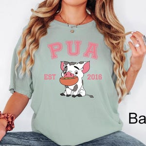 May include: A light green t-shirt featuring the word "PUA" in pink block letters above a cartoon pig holding a bowl of food. The text "EST 2016" is below the pig.