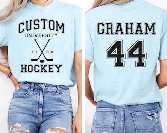 May include: Light blue t-shirt with "CUSTOM UNIVERSITY HOCKEY" text and crossed hockey sticks graphic on the front. The back of the shirt displays "GRAHAM 44" in black block letters. The shirt is paired with blue jeans.