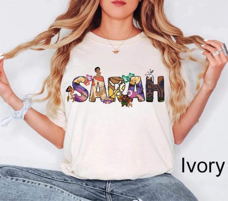 May include: Ivory-colored t-shirt featuring the name "SARAH" in colorful, illustrated letters. Each letter is decorated with characters and imagery from a popular animated film. The shirt is a casual, everyday wear item.