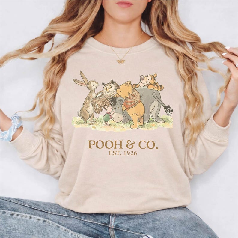 Puede incluir: Sudadera beige de cuello redondo con un dibujo de Winnie the Pooh y sus amigos. La ilustraci&oacute;n incluye un conejo, un b&uacute;ho, un burro, Winnie the Pooh y Tigger. El texto "POOH & CO. EST. 1926" est&aacute; impreso debajo.