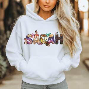 May include: White hooded sweatshirt with the name "SARAH" in colorful, decorative lettering. The letters are filled with various cartoon characters and floral designs, creating a playful and personalized aesthetic.