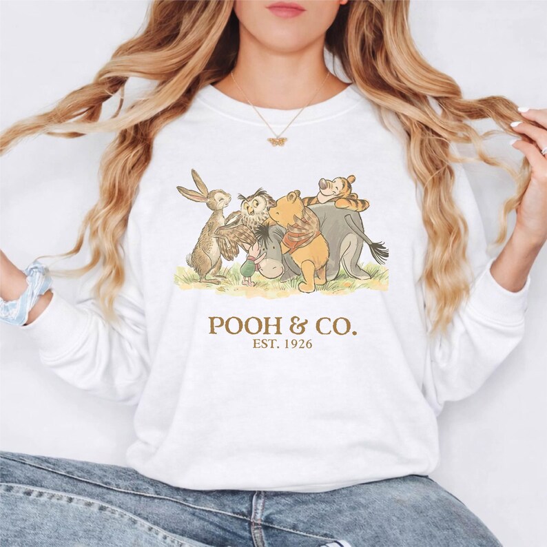 Puede incluir: Sudadera blanca de manga larga con un dibujo de Winnie the Pooh y sus amigos. La ilustraci&oacute;n incluye un conejo, un b&uacute;ho, un burro, un oso y un tigre. El texto "POOH & CO. EST. 1926" est&aacute; impreso debajo.