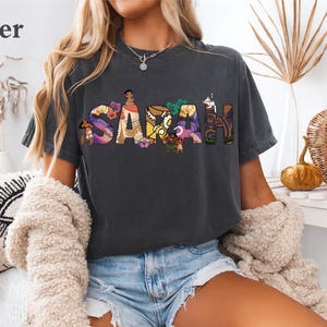 May include: Dark gray t-shirt with the name "SARAH" in colorful, illustrated letters. The letters are filled with images of cartoon characters. The shirt is worn with denim shorts and a cream-colored cardigan.