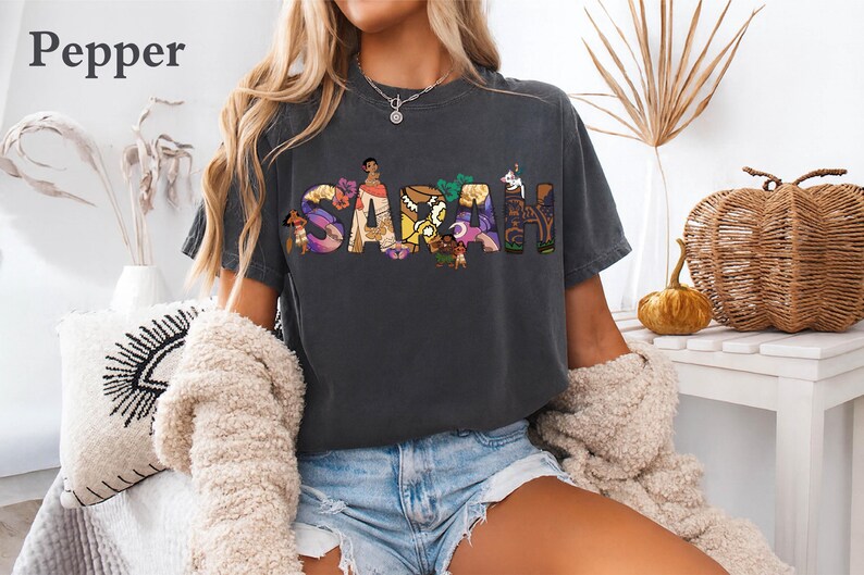 May include: Dark gray t-shirt with the name "SARAH" in colorful, illustrated letters. The letters are filled with images of cartoon characters. The shirt is worn with denim shorts and a cream-colored cardigan.