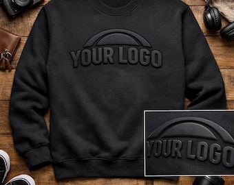 Puede incluir: Una sudadera negra con cuello redondo y un logotipo en relieve 3D "YOUR LOGO". La sudadera tiene un logotipo en relieve de primera calidad y un diseño en relieve 3D. La prenda se exhibe sobre una superficie de madera.
