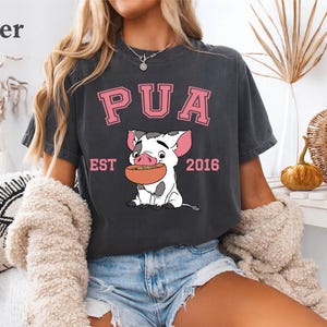 May include: Dark gray t-shirt featuring a cartoon pig holding a bowl of food. The shirt has the word "PUA" in pink block letters above the pig, with "EST 2016" below. The shirt is part of a clothing collection.