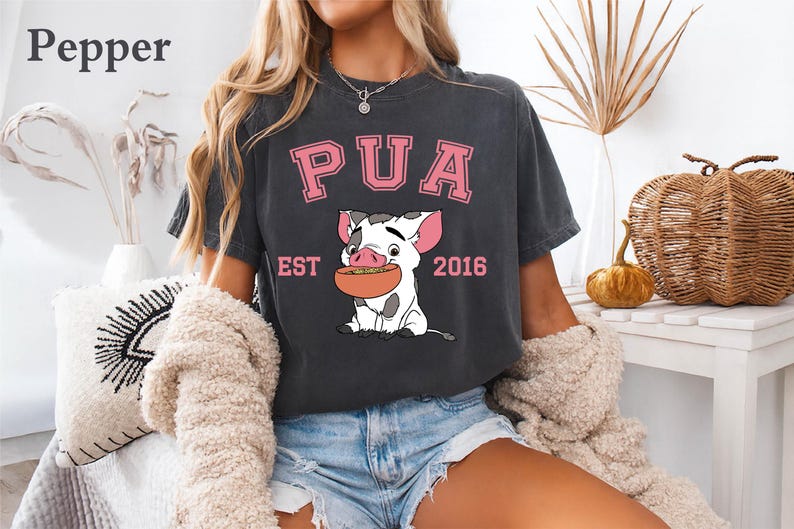May include: Dark gray t-shirt featuring a cartoon pig holding a bowl of food. The shirt has the word "PUA" in pink block letters above the pig, with "EST 2016" below. The shirt is part of a clothing collection.