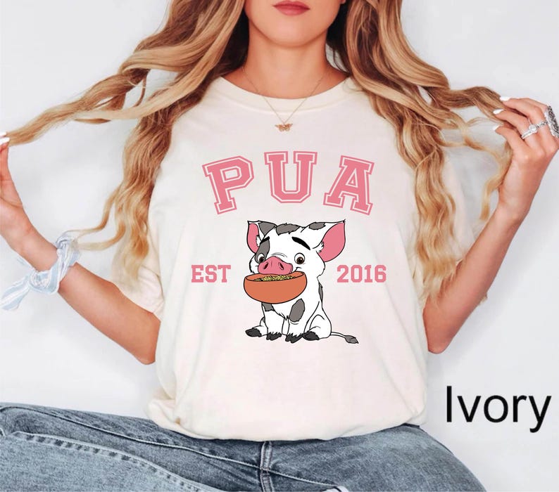 May include: Ivory-colored t-shirt featuring a cartoon pig holding a bowl of food. The word "PUA" is printed above the pig in pink block letters, with "EST 2016" below. The shirt is a casual style.