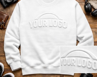 Puede incluir: Una sudadera blanca de cuello redondo con un diseño "YOUR LOGO" en relieve. La sudadera presenta un logotipo en relieve blanco de primera calidad y el texto "PREMIUM WHITE PUFF LOGO WHITE ON WHITE RAISED DESIGN".