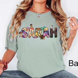 May include: A light green t-shirt with the name "SARAH" in colorful, floral-themed letters. The letters are decorated with cartoon characters. The word "Bay" is printed in black on the bottom right.