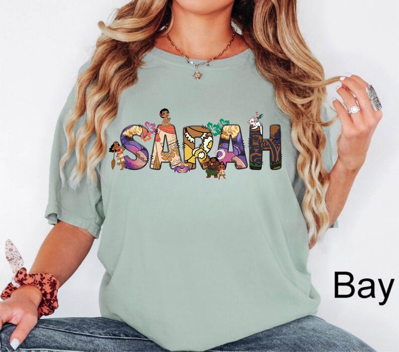 May include: A light green t-shirt with the name "SARAH" in colorful, floral-themed letters. The letters are decorated with cartoon characters. The word "Bay" is printed in black on the bottom right.