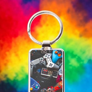 May include: Silver keychain with a rectangular pendant featuring a collage of colorful video game controllers. The background is a vibrant rainbow of colors. The keychain is a perfect accessory for gamers.