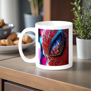 May include: White ceramic mug featuring a close-up of Spider-Man's mask. The design showcases the red and black webbed texture with a blue-tinted lens reflecting a cityscape. The mug has a curved handle and is set on a wooden surface.