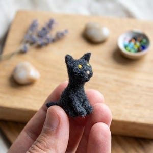 Peut inclure: Figurine de chat noir miniature aux yeux jaunes, fabriquée à partir d'un matériau gris foncé. Le chat est assis sur une surface en bois, avec des éléments décoratifs tels que des pierres et un petit bol de perles colorées.