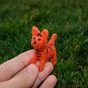 May include: A small, orange felted cat figurine with black stripes and facial details. The handmade cat is standing on a green grassy surface. The cat is a whimsical, handcrafted item.