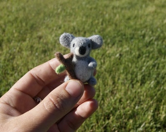 May include: A handcrafted grey felt koala bear figure, holding a brown branch with a green leaf. The koala has a black nose and small, rounded ears. The figure is held in a hand against a blurred green grass background.