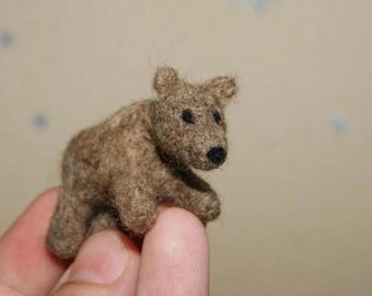 May include: A small, needle-felted brown bear figurine crafted from textured wool fibres. The bear features a rounded body, small ears, and two tiny black bead eyes with a matching black nose, showcasing a soft, fuzzy appearance throughout.