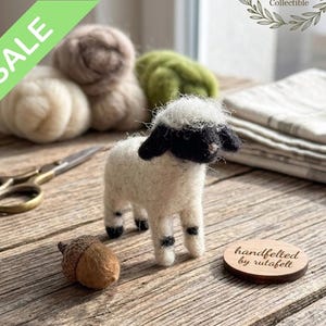 Op de afbeelding: Een handgemaakt vilten lammetjesfiguur met een zwart-witte kop en poten. Het woord "SALE" staat op een groene banner. Andere items zijn wol, een schaar en een eikel.