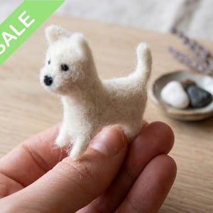 Puede incluir: Una pequeña figura de perro de fieltro blanco con ojos y nariz negros. El perro está de pie sobre sus cuatro patas. Una pancarta verde en la esquina superior izquierda dice "SALE". La figura se sostiene en una mano.