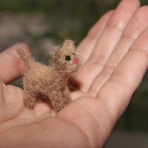 May include: A miniature, handmade cat figurine crafted from tan wool. The tiny feline has black eyes and a pink nose, standing on four legs with a small tail. The cat is held in an open hand, showcasing its small size.
