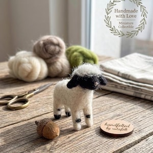 May include: A miniature felted lamb with a white body, black head and legs, and a fluffy white top. The lamb is on a wooden surface next to an acorn, wool, and a wooden tag that says "handfelted by rutafelt". The image also includes the text "Handmade with Love".
