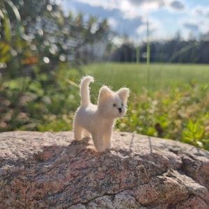 May include: A miniature white felt cat figurine, with small ears and a curled tail, stands on a large, textured rock. The background shows green grass, foliage, and a bright sky, creating a natural outdoor setting.