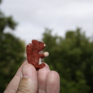 May include: A small, hand-crafted felt squirrel figurine in shades of brown and white. The squirrel is posed standing on its hind legs with its tail curled up.