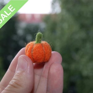 May include: A miniature orange felt pumpkin with a green stalk. The pumpkin is held in a hand against a blurred green backdrop. A green banner in the upper left corner reads "SALE".