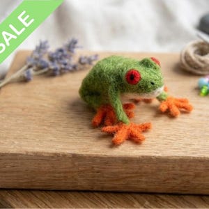 May include: A handmade green felt frog with red eyes and orange feet. The frog is displayed on a wooden surface, with a green banner displaying the word "SALE" in the upper left corner. A charming decorative item.