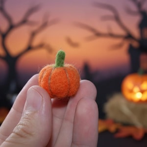 Könnte beinhalten: Ein kleiner, orangefarbener Filzkürbis mit grünem Stiel wird in einer Hand gehalten. Der Hintergrund ist verschwommen, aber mit einem beleuchteten Jack-o'-Lantern, Bäumen und einem gruseligen Haus, passend zum Halloween-Thema.