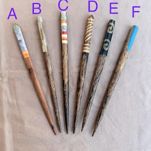 May include: Six wooden hair sticks labeled A through F. Stick A has a multi-colored inlay, B has a light inlay, C features horizontal stripes, D has white diagonal lines, E has gold swirl patterns on black, and F has a blue inlay at the top.