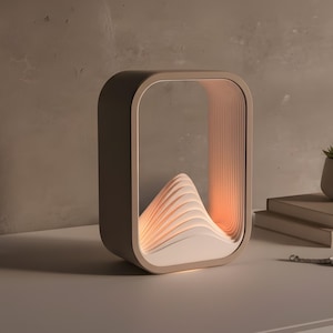 Puede incluir: Una l&aacute;mpara de mesa moderna con luz c&aacute;lida. Presenta un dise&ntilde;o en capas en forma de monta&ntilde;a dentro de un marco neutro. Una peque&ntilde;a suculenta en maceta blanca, libros y una llave est&aacute;n sobre la mesa.
