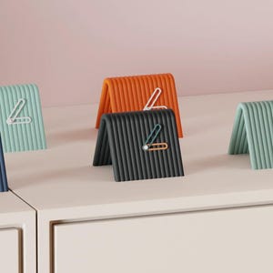 Op de afbeelding: Vijf kleine, tentvormige analoge klokken met geribbelde oppervlakken in marineblauw, lichtgroen, oranje en zwart. Elke klok heeft een eenvoudig wijzerplaatmechanisme met twee wijzers in een contrasterende kleur op de voorzijde.