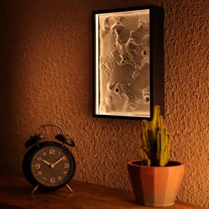 Könnte beinhalten: Ein schwarz gerahmtes Wandkunstwerk mit einer strukturierten, mondähnlichen Oberfläche, von hinten beleuchtet. Ein schwarzer Wecker und ein Topfkaktus stehen auf einer Holzoberfläche unter dem Kunstwerk. Die Wand ist strukturiert und braun.