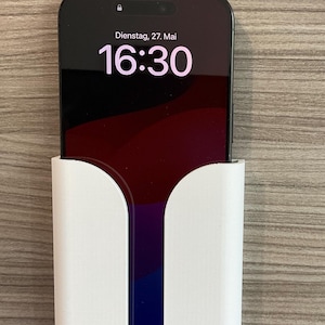 Może przedstawiać: Czarny smartfon wyświetlający godzinę 16:30 na czerwono-fioletowym tle, trzymany w białym uchwycie. Na ekranie wyświetlana jest również data i dzień tygodnia. Telefon jest podłączony do białego kabla ładującego.