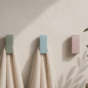 Peut inclure: Trois patères murales rectangulaires de couleur vert sauge, bleu clair et rose pâle, chacune présentant une surface texturée distincte. Deux des patères soutiennent des serviettes beiges pliées, tandis que la troisième reste vide sur le mur.