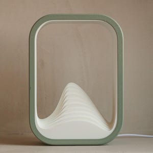 Puede incluir: L&aacute;mpara de mesa moderna y minimalista con un marco rectangular verde salvia. En el interior, una estructura blanca en forma de ola proporciona la fuente de luz. Un cable blanco se extiende desde la base. L&aacute;mpara decorativa.