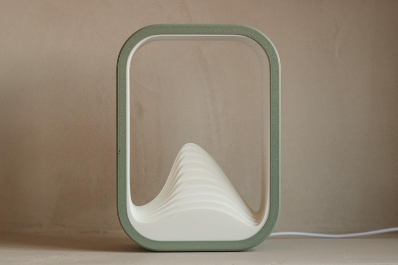Puede incluir: L&aacute;mpara de mesa moderna y minimalista con un marco rectangular verde salvia. En el interior, una estructura blanca en forma de ola proporciona la fuente de luz. Un cable blanco se extiende desde la base. L&aacute;mpara decorativa.