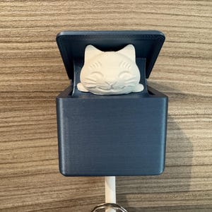 Könnte beinhalten: Ein marineblauer Schlüsselhalter mit einer weißen Katzenfigur im Inneren. Der rechteckige Schlüsselhalter hat einen Klappdeckel. Ein weißer Stift ragt von unten heraus und hält einen silbernen Schlüsselring.