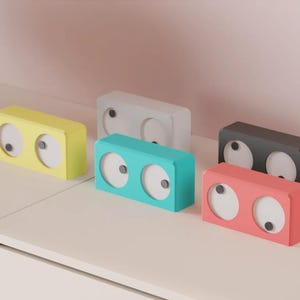 Può includere: Cinque orologi rettangolari in giallo, grigio chiaro, turchese, grigio scuro e corallo. Ogni orologio presenta una cornice a tinta unita con due quadranti bianchi circolari, ciascuno dotato di una piccola manopola grigio scuro al centro.