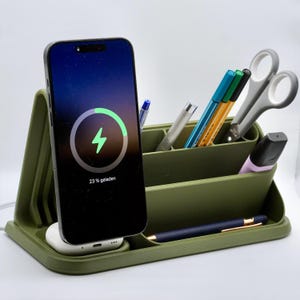 Könnte beinhalten: Olivgrüner Schreibtisch-Organizer mit einem Smartphone, das geladen wird. Das Telefon zeigt ein Ladesymbol und den Text "23% geladen". Der Organizer enthält Stifte, eine Schere und einen Textmarker. Ein weißes Ladepad ist sichtbar.