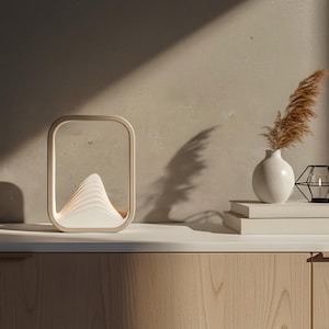 Puede incluir: Una l&aacute;mpara de mesa moderna y rectangular con un dise&ntilde;o en forma de monta&ntilde;a, que emite una luz suave y c&aacute;lida. La l&aacute;mpara es de color beige y est&aacute; sobre una superficie de madera. Un jarr&oacute;n blanco con hierba seca y un portavelas geom&eacute;trico negro tambi&eacute;n est&aacute;n presentes.