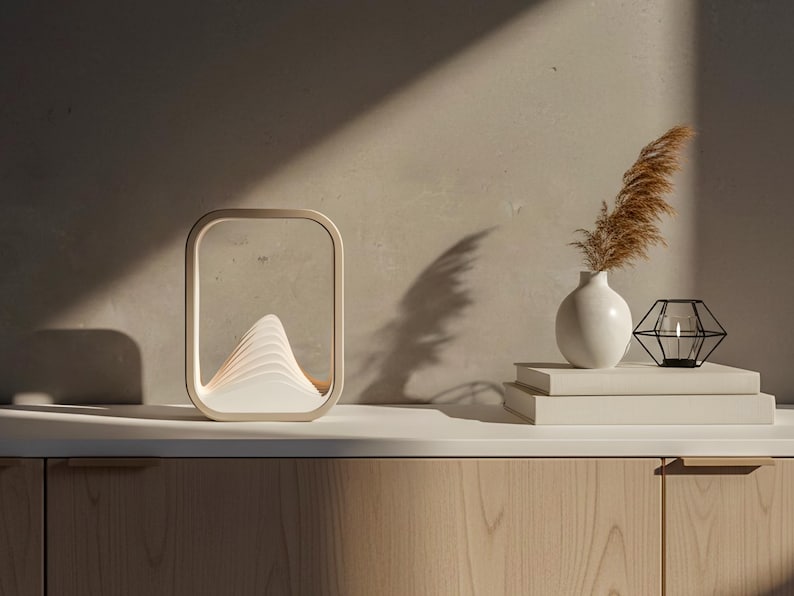 Puede incluir: Una l&aacute;mpara de mesa moderna y rectangular con un dise&ntilde;o en forma de monta&ntilde;a, que emite una luz suave y c&aacute;lida. La l&aacute;mpara es de color beige y est&aacute; sobre una superficie de madera. Un jarr&oacute;n blanco con hierba seca y un portavelas geom&eacute;trico negro tambi&eacute;n est&aacute;n presentes.