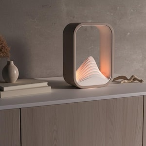 Puede incluir: L&aacute;mpara moderna de tonos neutros con marco rectangular y dise&ntilde;o en capas en forma de monta&ntilde;a en el interior. La l&aacute;mpara est&aacute; sobre una superficie blanca con un jarr&oacute;n, libros y una escultura de madera. Mide aproximadamente 30 cm de alto y 20 cm de ancho.
