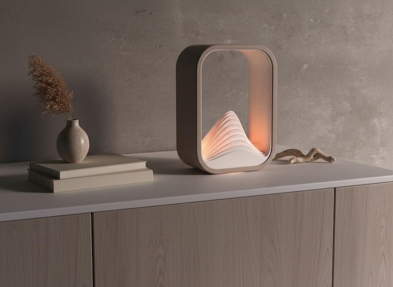 Puede incluir: L&aacute;mpara moderna de tonos neutros con marco rectangular y dise&ntilde;o en capas en forma de monta&ntilde;a en el interior. La l&aacute;mpara est&aacute; sobre una superficie blanca con un jarr&oacute;n, libros y una escultura de madera. Mide aproximadamente 30 cm de alto y 20 cm de ancho.