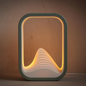 Puede incluir: Una l&aacute;mpara de mesa rectangular moderna con un marco verde salvia. La l&aacute;mpara presenta un dise&ntilde;o en capas en forma de ola en el centro, iluminado por una luz amarilla c&aacute;lida. La l&aacute;mpara est&aacute; enchufada con un cable blanco.