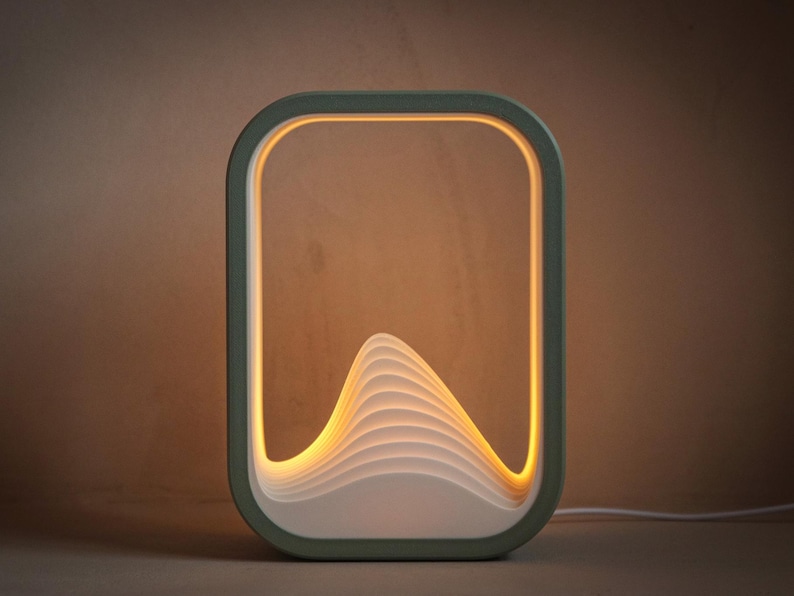 Puede incluir: Una l&aacute;mpara de mesa rectangular moderna con un marco verde salvia. La l&aacute;mpara presenta un dise&ntilde;o en capas en forma de ola en el centro, iluminado por una luz amarilla c&aacute;lida. La l&aacute;mpara est&aacute; enchufada con un cable blanco.