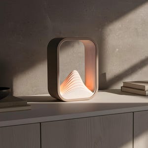 Puede incluir: L&aacute;mpara de mesa contempor&aacute;nea con un dise&ntilde;o &uacute;nico. La l&aacute;mpara tiene un marco rectangular con una estructura interna en capas que evoca una monta&ntilde;a, emitiendo una luz c&aacute;lida. Se encuentra sobre una superficie blanca, con un cuenco oscuro y libros. Aproximadamente 20 cm.