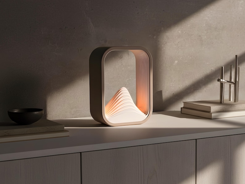 Puede incluir: L&aacute;mpara de mesa contempor&aacute;nea con un dise&ntilde;o &uacute;nico. La l&aacute;mpara tiene un marco rectangular con una estructura interna en capas que evoca una monta&ntilde;a, emitiendo una luz c&aacute;lida. Se encuentra sobre una superficie blanca, con un cuenco oscuro y libros. Aproximadamente 20 cm.